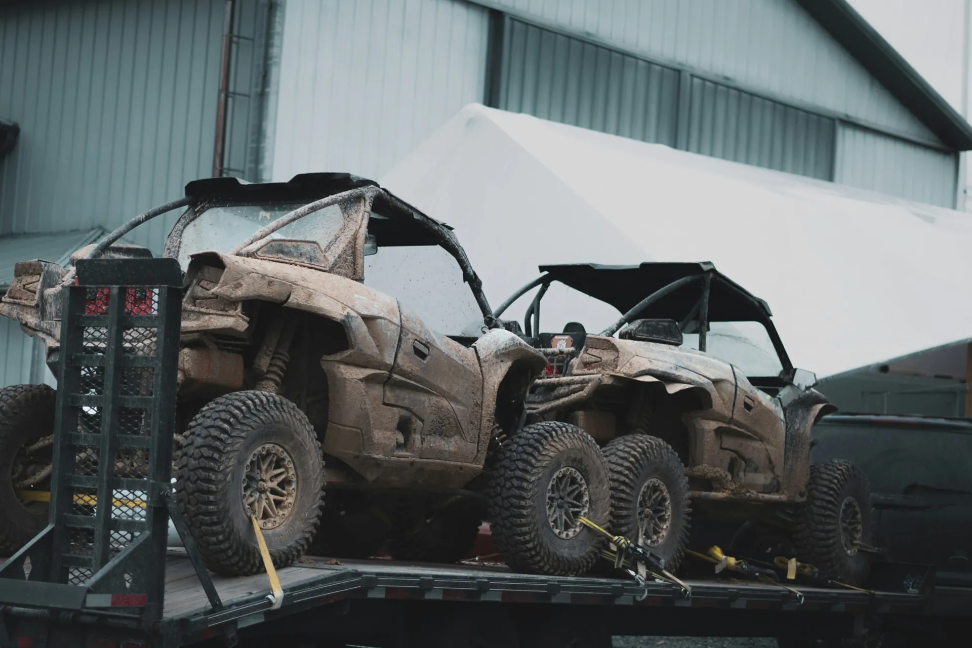 Two mud-covered UTVs strapped to a trailer outside a shop building